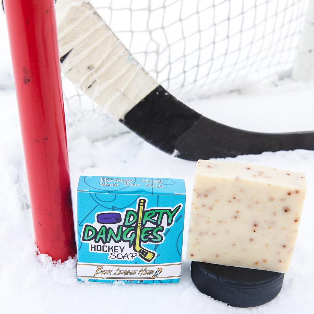 A black soap bar sits in the snow with a hockey goal, hockey puck and a hockey stick. Puck Drop