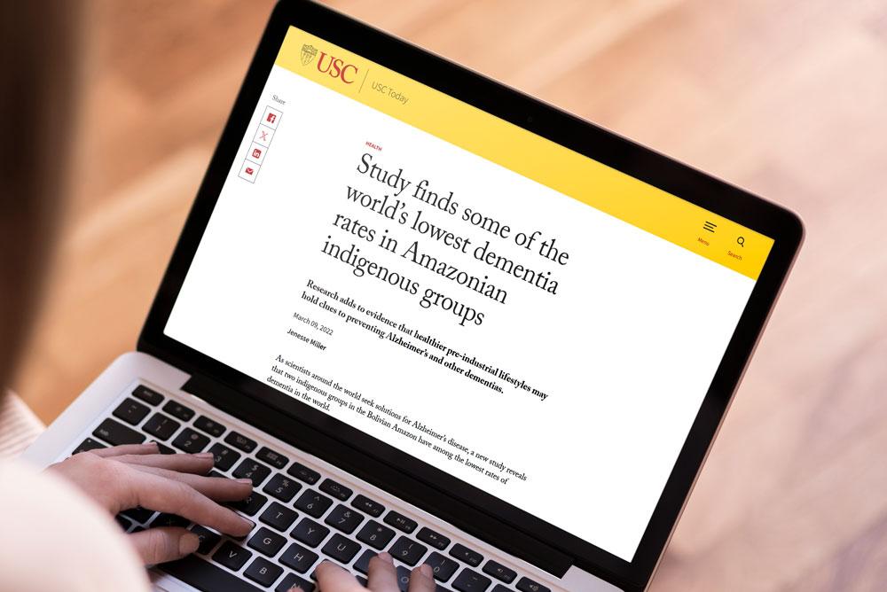 Person typing on a laptop displaying a USC news article titled “Study finds some of the world’s lowest dementia rates in Amazonian indigenous groups.”