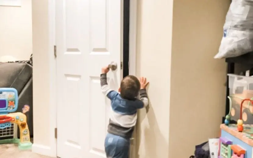 Toddler reaching up to turn a door handle and open a closed door, illustrating how easily a toddler can access off limits areas like a room with a litter box without proper childproofing.