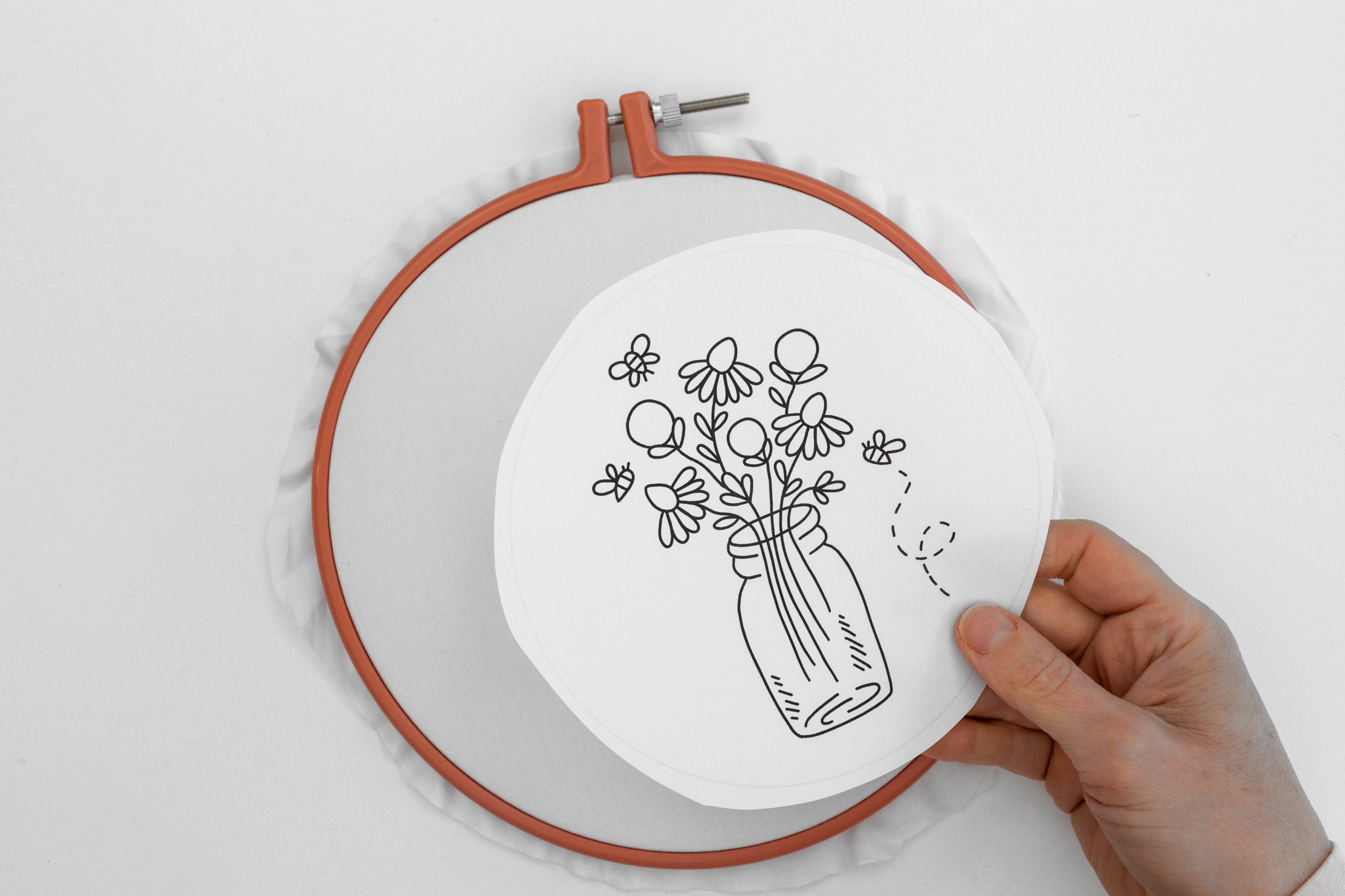 A hand holds a floral vase pattern on top of a blank hoop.