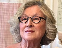 Portrait of a graceful 70-year-old woman wearing stylish glasses, her complexion radiant and youthful, likely due to Bella Terra Oils, set against a cozy bathroom interior with pink checkered curtains.