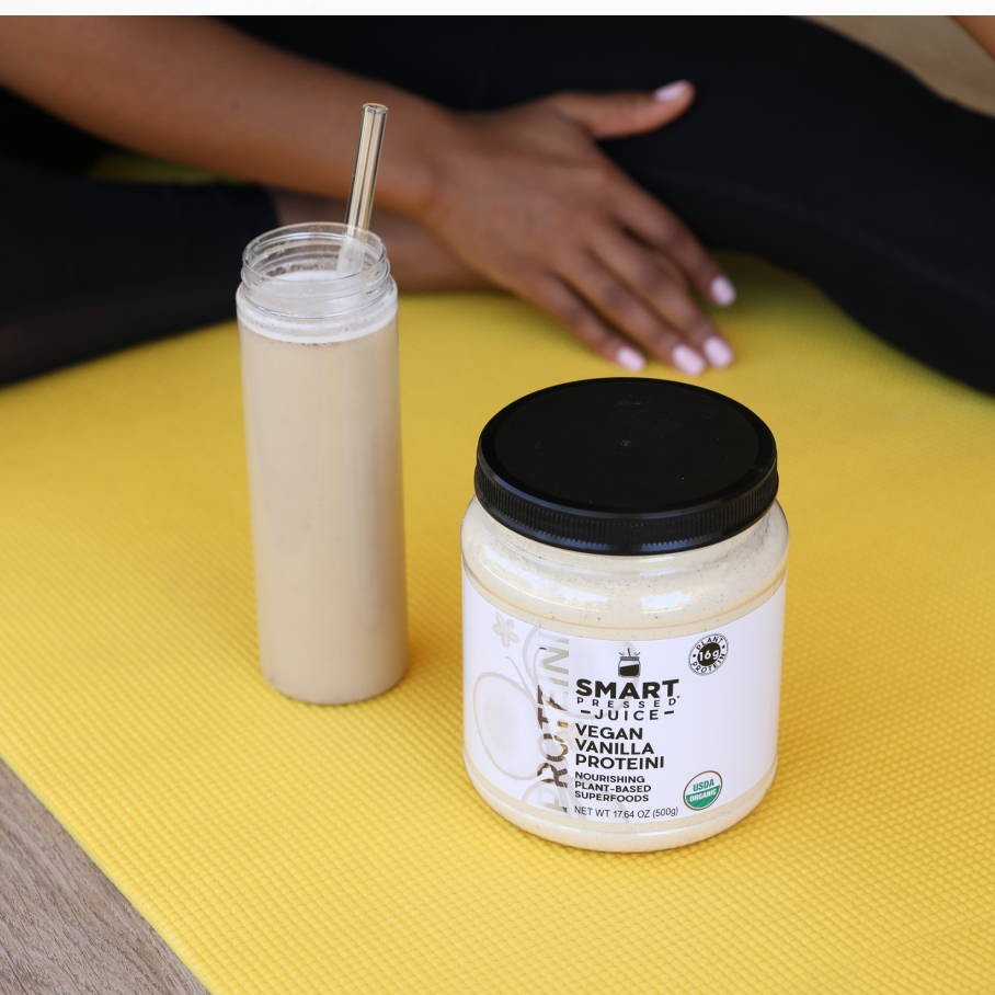 A tall glass of white smoothie with a straw beside a jar of Vegan Vanilla Proteini set on a yellow mat.