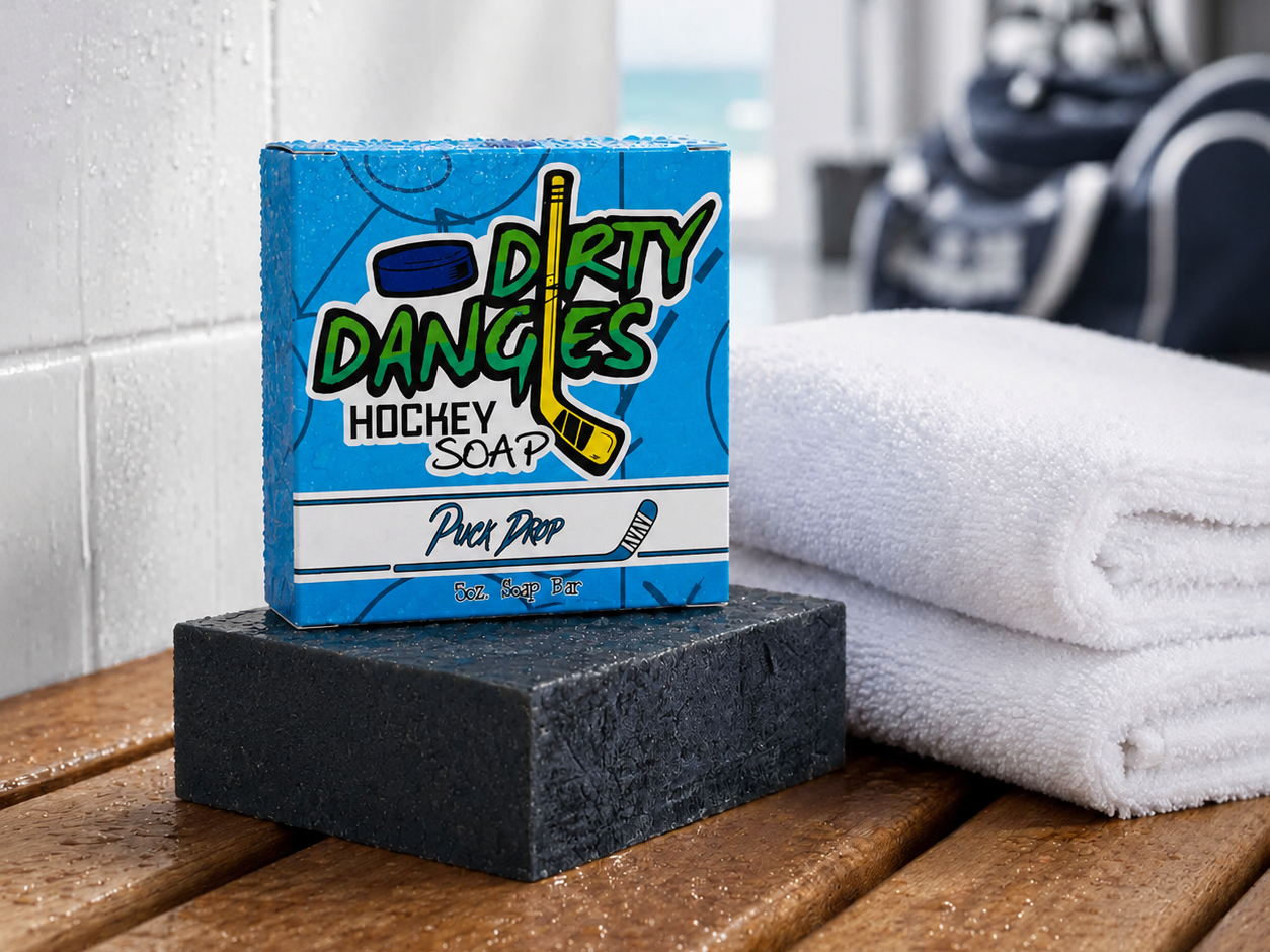 Puck Drop hockey soap bar and box on a wet locker room bench beside folded white towels with hockey gear in the background.