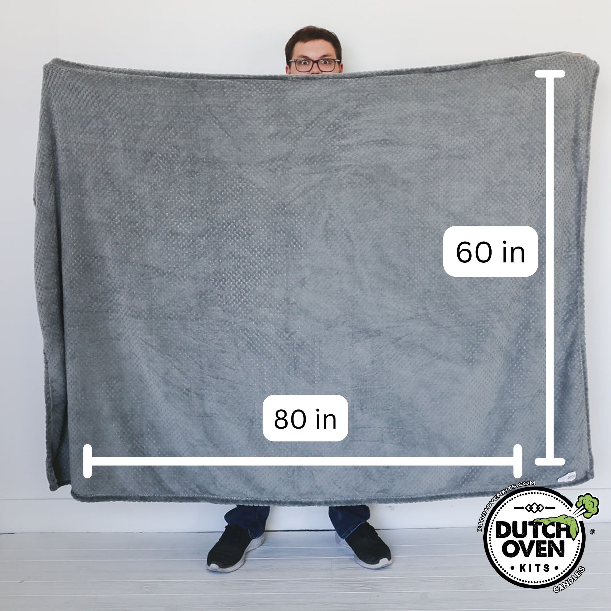A man holding up a gray dutch oven kits fart blanket displaying it's size. 60in x 80in