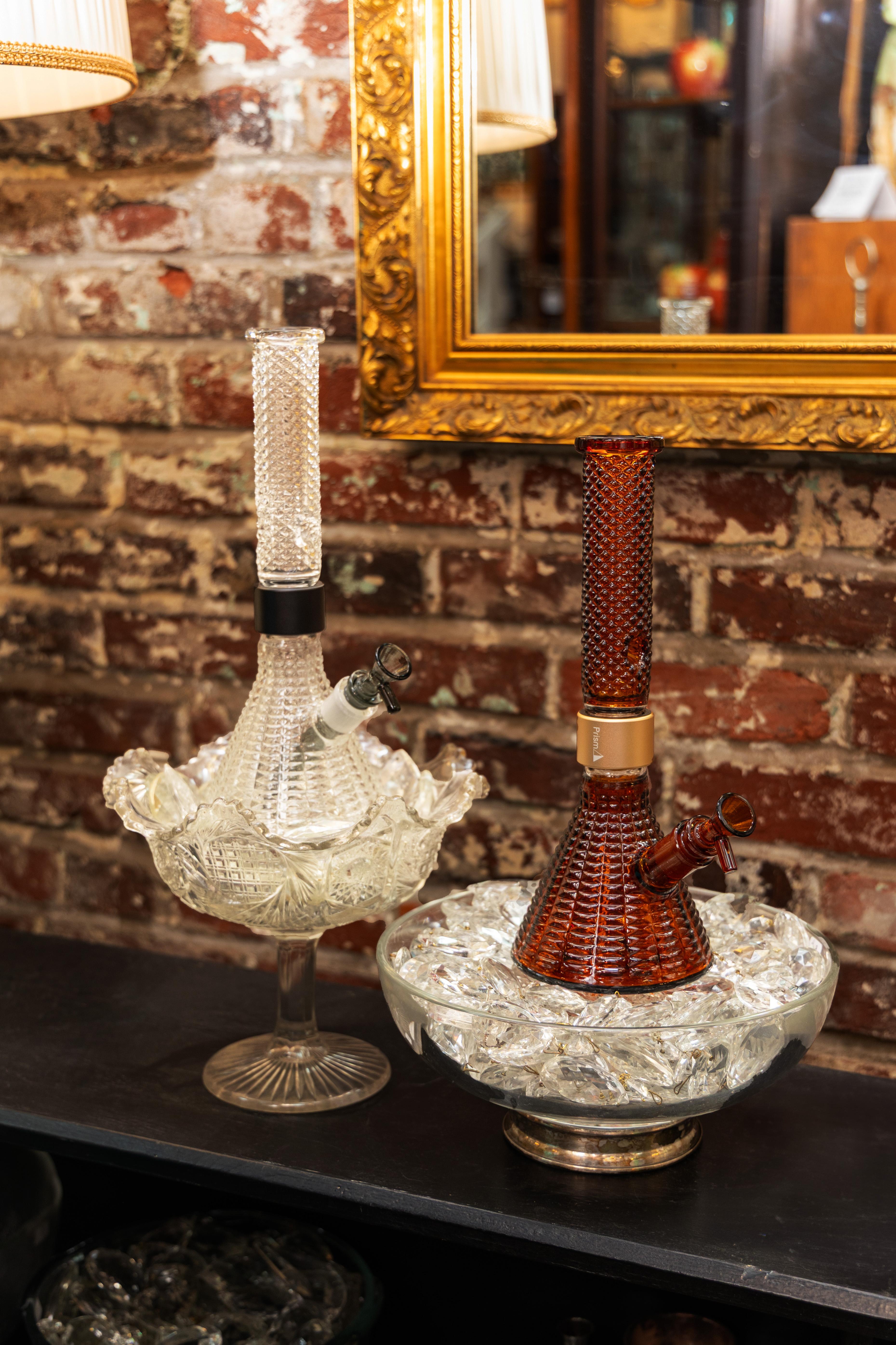 The Prism x Jane West Twenties MINI Beaker Bongs, in clear and vintage amber, showcased as elegant centerpieces within antique party ice bowls. The photo emphasizes how the Art Deco diamond texture and the piece's design allow it to be seamlessly integrated into classy, sophisticated cannabis consumption events.