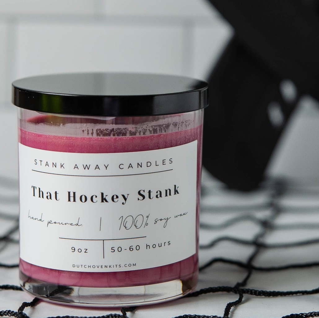 picture of a maroon wine cellar scent candle sitting on a hockey net with hockey sticks in the background. That Hockey Stank