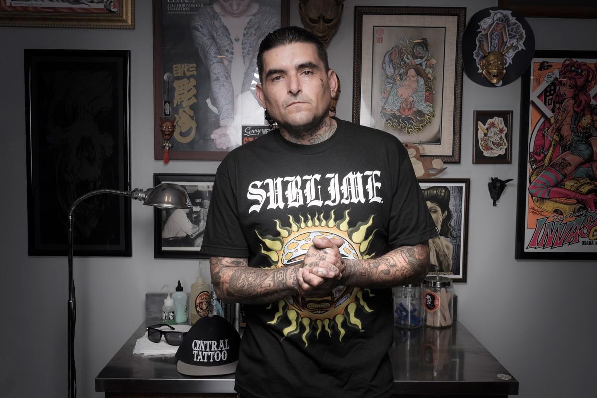 Legendary artist Opie Ortiz in his tattoo studio wearing a Sublime Sun logo t-shirt with Gothic font. The image showcases the original 40oz. to Freedom sun artwork and tattoo lettering that inspired the decals on the Prism x Sublime modular glass collection.