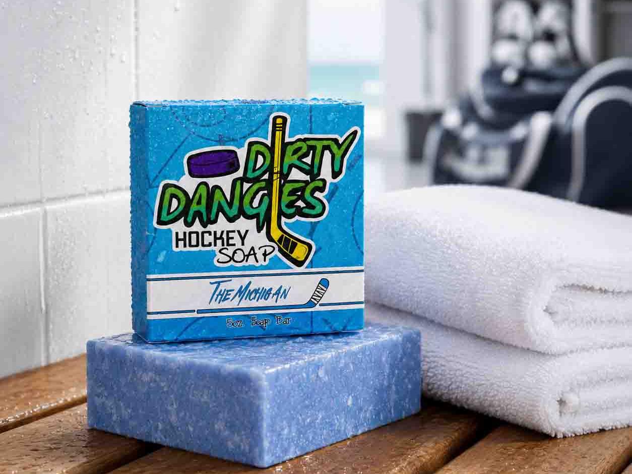 The Michigan hockey soap bar and box on a wet locker room bench beside folded white towels with hockey gear in the background.
