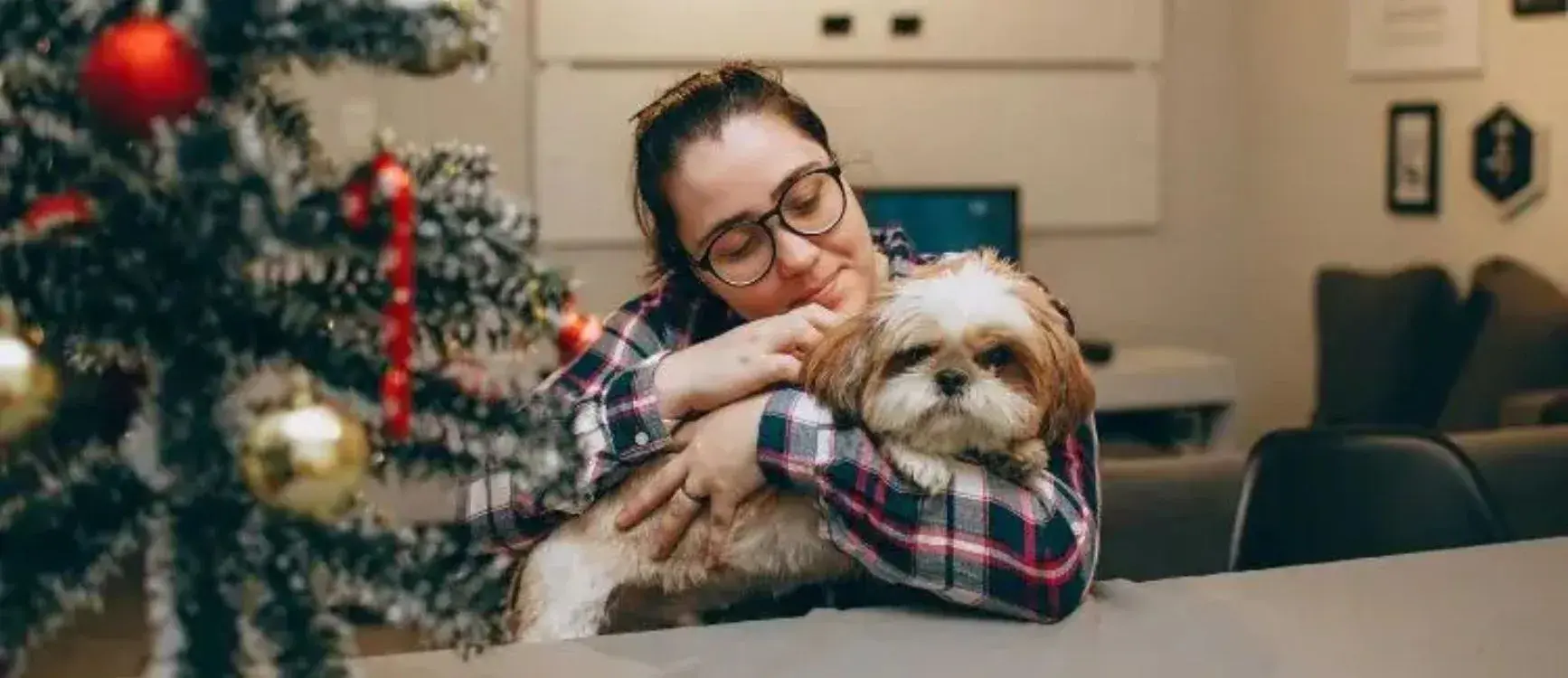 A woman embraces her dog in front of a beautifully decorated Christmas tree, radiating warmth and holiday spirit.