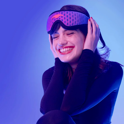 A smiling girl holding a blue mesh sleep mask to her head.