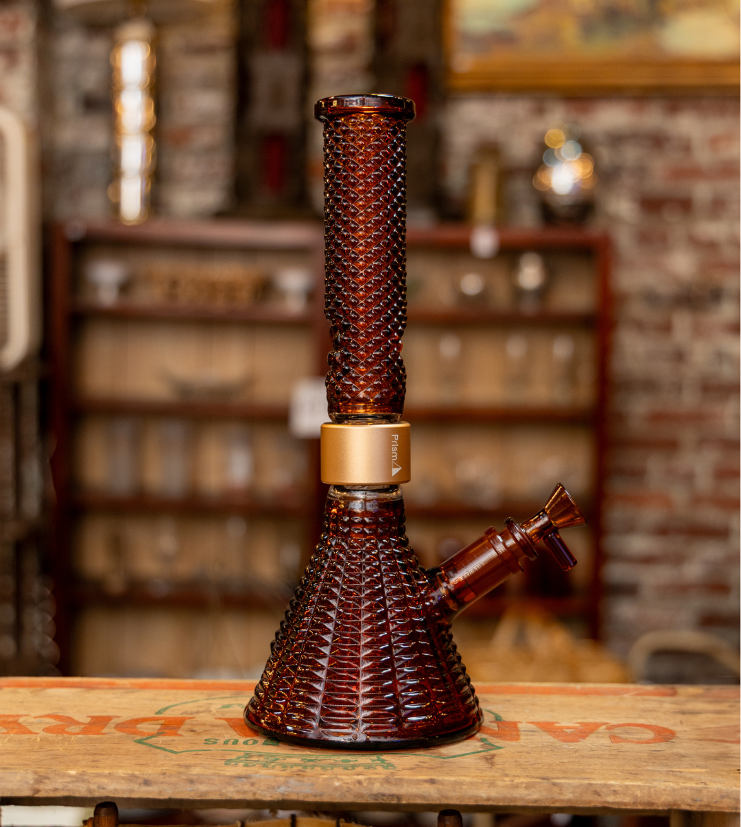 The Prism x Jane West Twenties MINI Beaker Bong in the exclusive vintage amber colorway, placed on an antique cigar box. The warm, inviting setting, featuring red brick walls and a fine china cabinet, emphasizes the bong's role as a luxurious, collector's piece and a sophisticated accessory for the modern host.