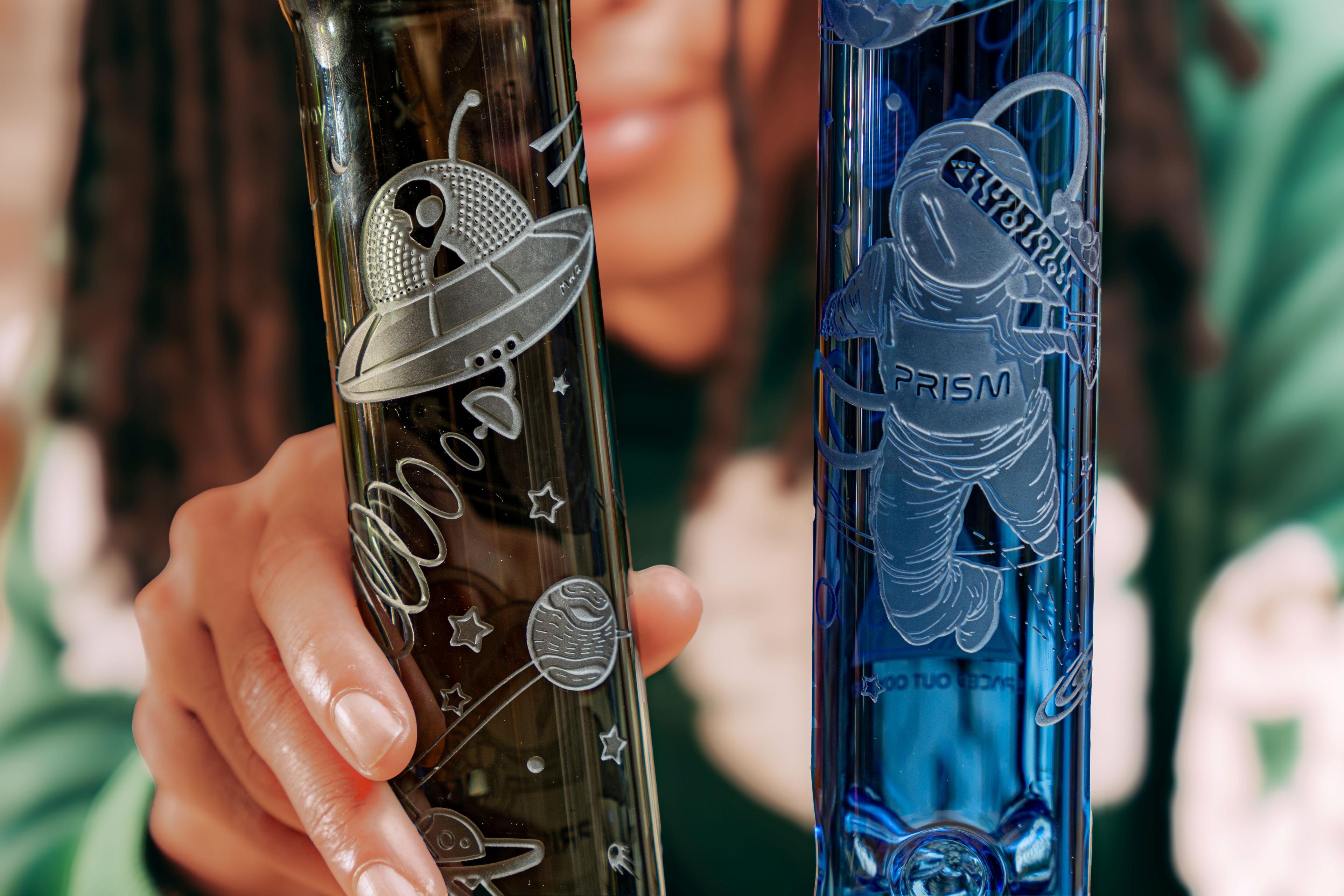 A lifestyle photo of a person holding two Milkyway x Prism spaced out Odyssey etched glass bong mouthpieces, one sapphire and one midnight colored