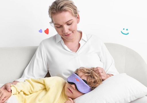 A little boy wearing a purple contoured sleep mask for kids, lying down on a blonde woman’s lap.