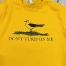 Close up of a gold and yellow t shirt with a picture of a seagull standing in the grass. Text on the shirt says "Don't Turd On Me." Similar to the Gadsden "Dont Tread On Me" Flag