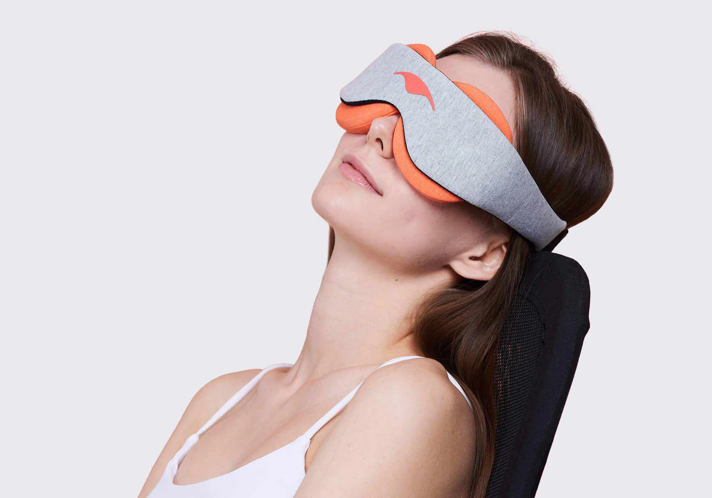 A girl leaning back against a black chair, wearing a light gray sleep mask with orange warming eye cups.