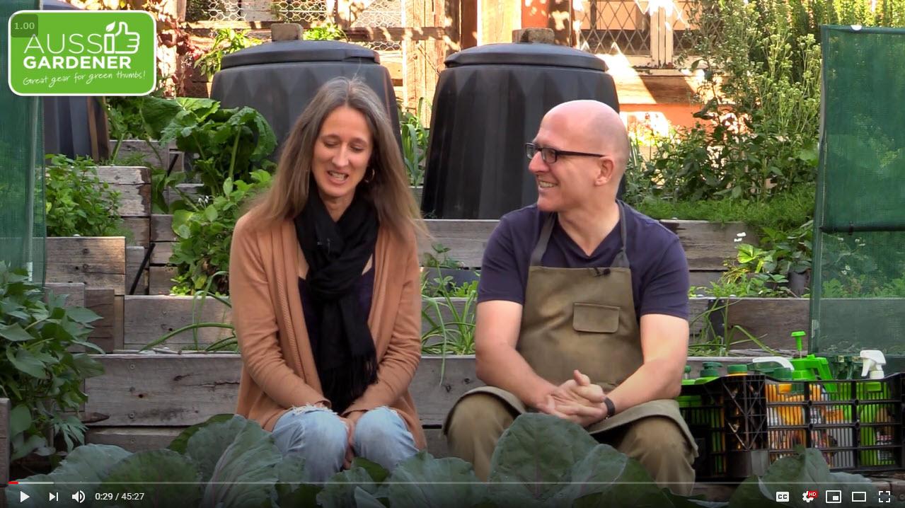 Brian and Kaylene sitting in a vegetable garden, talking and smiling during a gardening video.