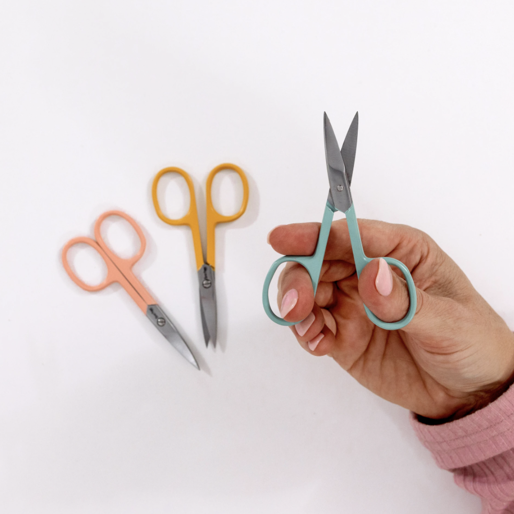 This is an image of a hand holds an embroidery scissor with other scissors in the background, available for purchase from the Clever Poppy Shop.