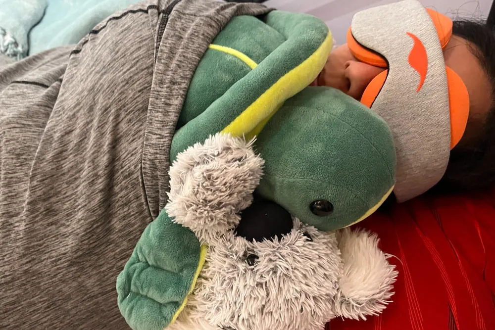 A woman lying down with a turtle stuffed toy, wearing a gray sleep mask with heated eye cups.