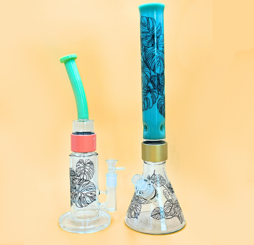 Two Prism modular bongs featuring the Monstera Decal: A Key Lime Green Bent Mouthpiece Big Honeycomb Bong with a coral connector, alongside a Tall Mint Green Mouthpiece Beaker Bong. The side-by-side display demonstrates the decal's aesthetic fit on both advanced percolation bases and standard beaker setups.