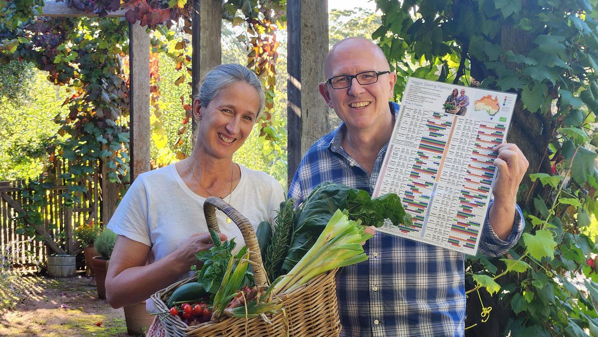 Brian and Kaylene standing in a leafy garden under a vine-covered pergola holding a colourful Veggie Sowing Planner chart and a wicker basket filled with freshly harvested vegetables, including leafy greens, leeks, herbs and tomatoes.