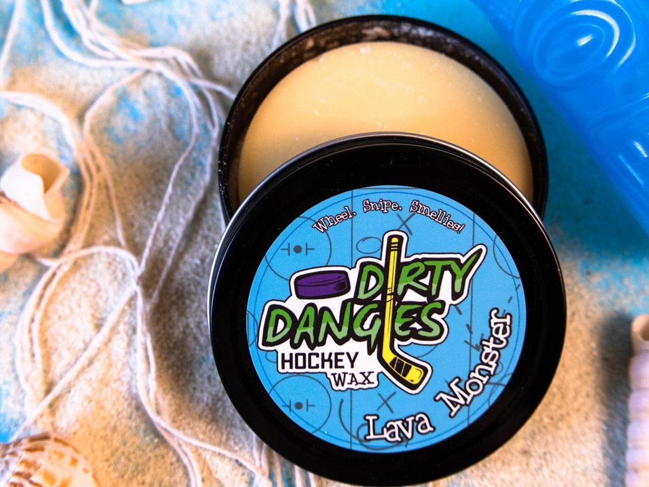 A tin of Dirty Dangles Hockey Stick Wax Lava Monster Scent sits open on a blue background surrounded by sand, seashells and a tiki mug