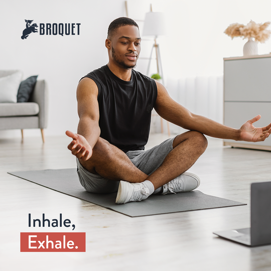 man in a yoga pose, sitting on a yoga mat on the floor, looking at a laptop, broquet logo, text reads: Inhale, exhale.
