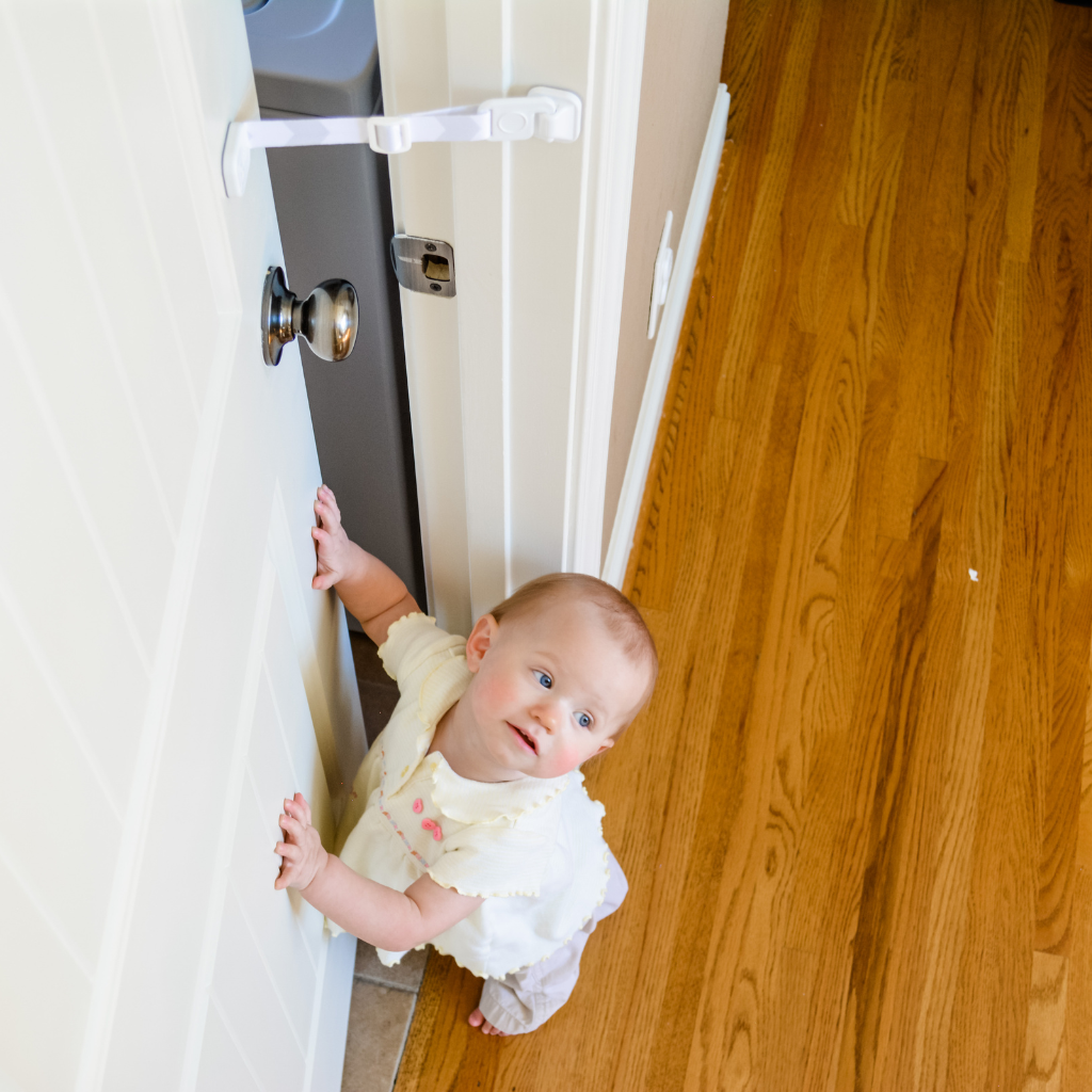 A baby stands behind a partially open door secured with The Door Buddy door strap. The adjustable strap baby proofs the room by keeping the door slightly open for cats while helping prevent babies and toddlers from entering.