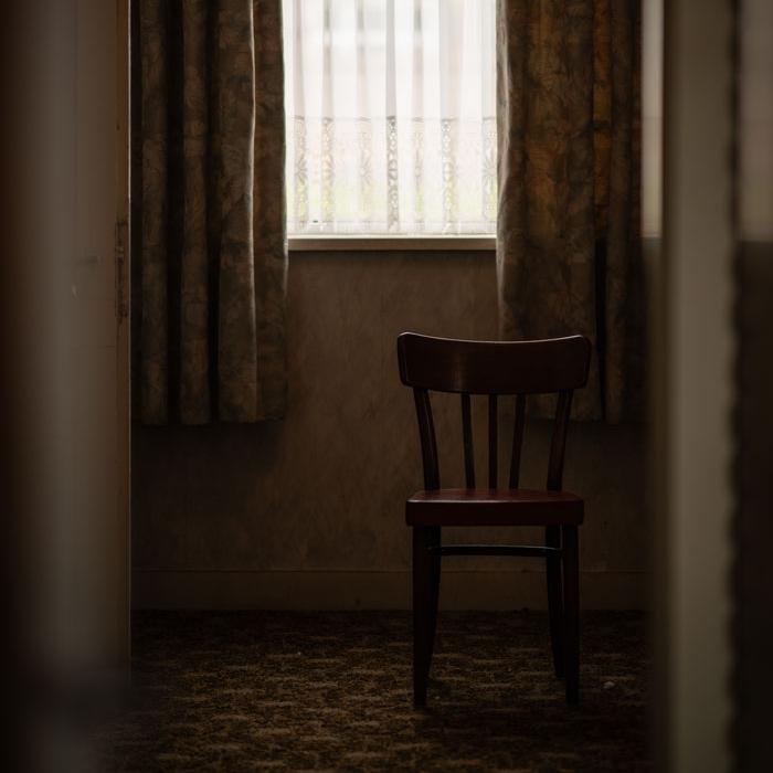 Empty wooden chair placed in a dimly lit room near a curtained window, evoking a quiet or lonely atmosphere.