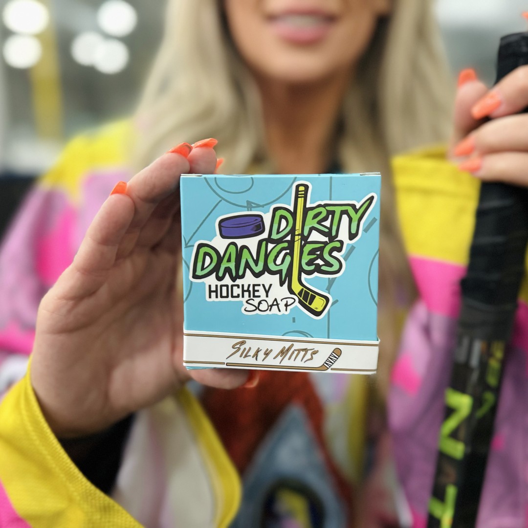 a woman hockey player holding a blue box of dirty dangles hockey soap silky mitts scent inside an ice rink.
