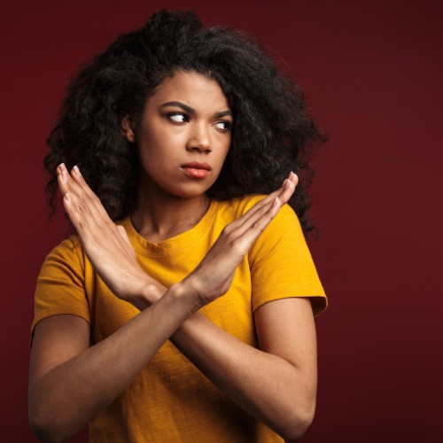 deep red background. young woman wearing a mustard yellow shirt holding up her arms in an "x" as if to say no