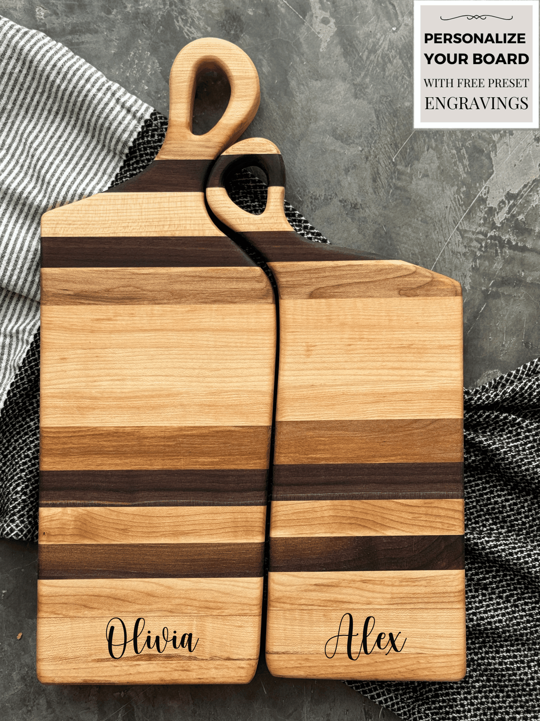 Two Nestling handcrafted wooden charcuterie boards made from maple and walnut with contrasting dark and light stripes. Both feature ergonomic handles with a soft, rounded design, displayed on a textured fabric backdrop for a rustic aesthetic.