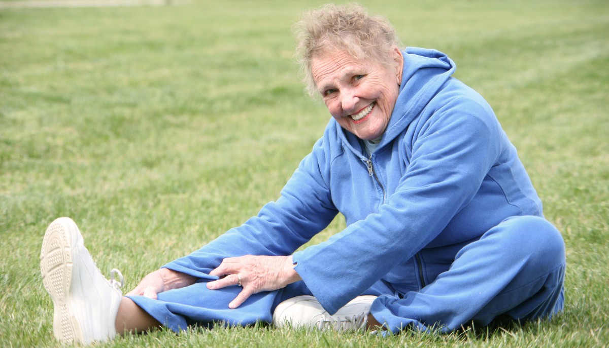 older woman wearing a blue sweats outfit, smiling and stretching her hamstring while sitting in the green grass.