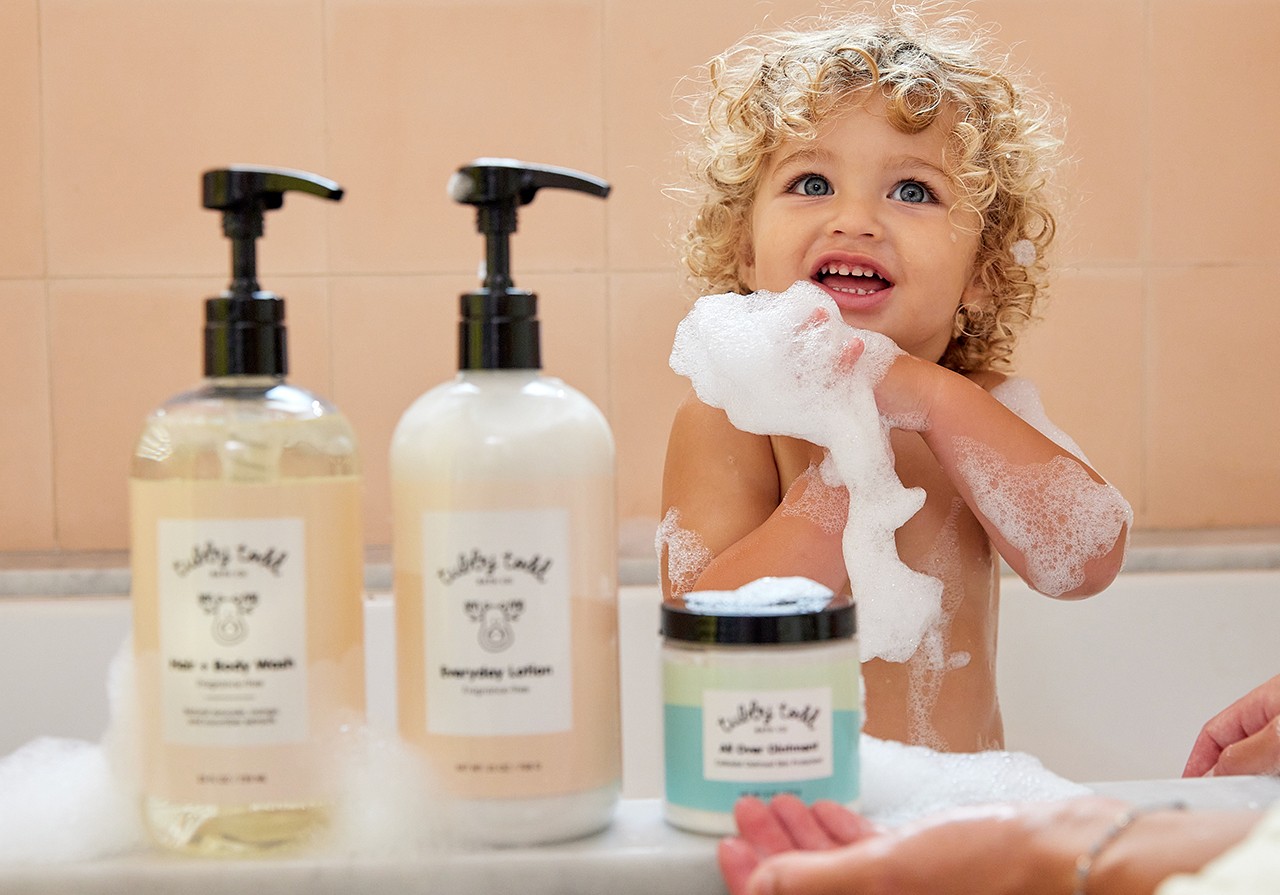 The Fragrance Free Regulars Bundle products are on the bath tub and the baby is standing behind them.