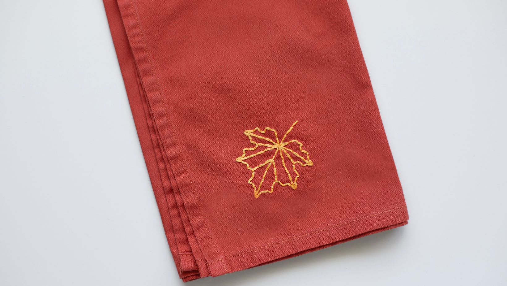 A yellow maple leaf outline embroidered on the corner of a rust-colored napkin.