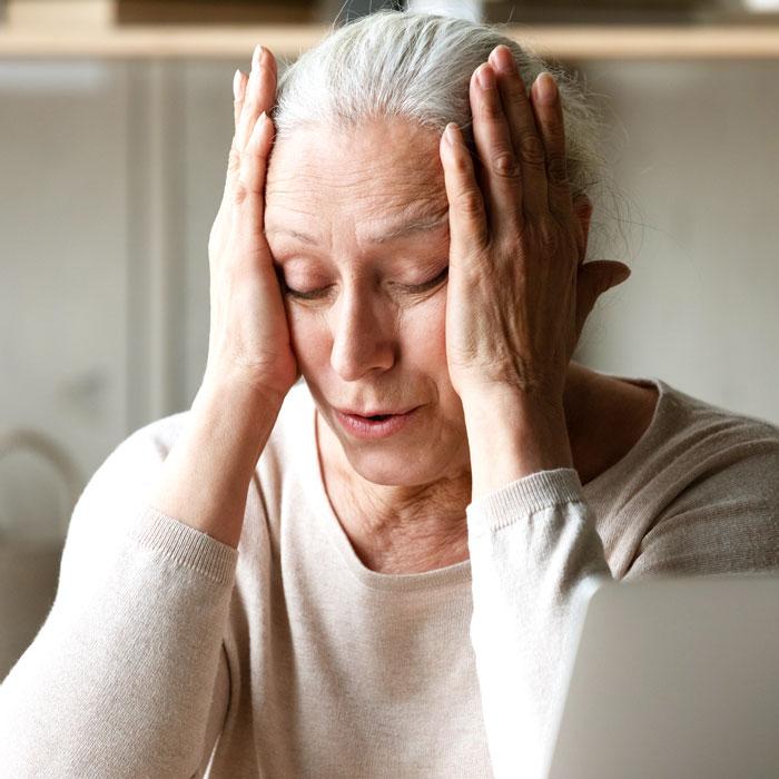 Elderly woman sitting with eyes closed and hands pressed to her temples, appearing tired or stressed.
