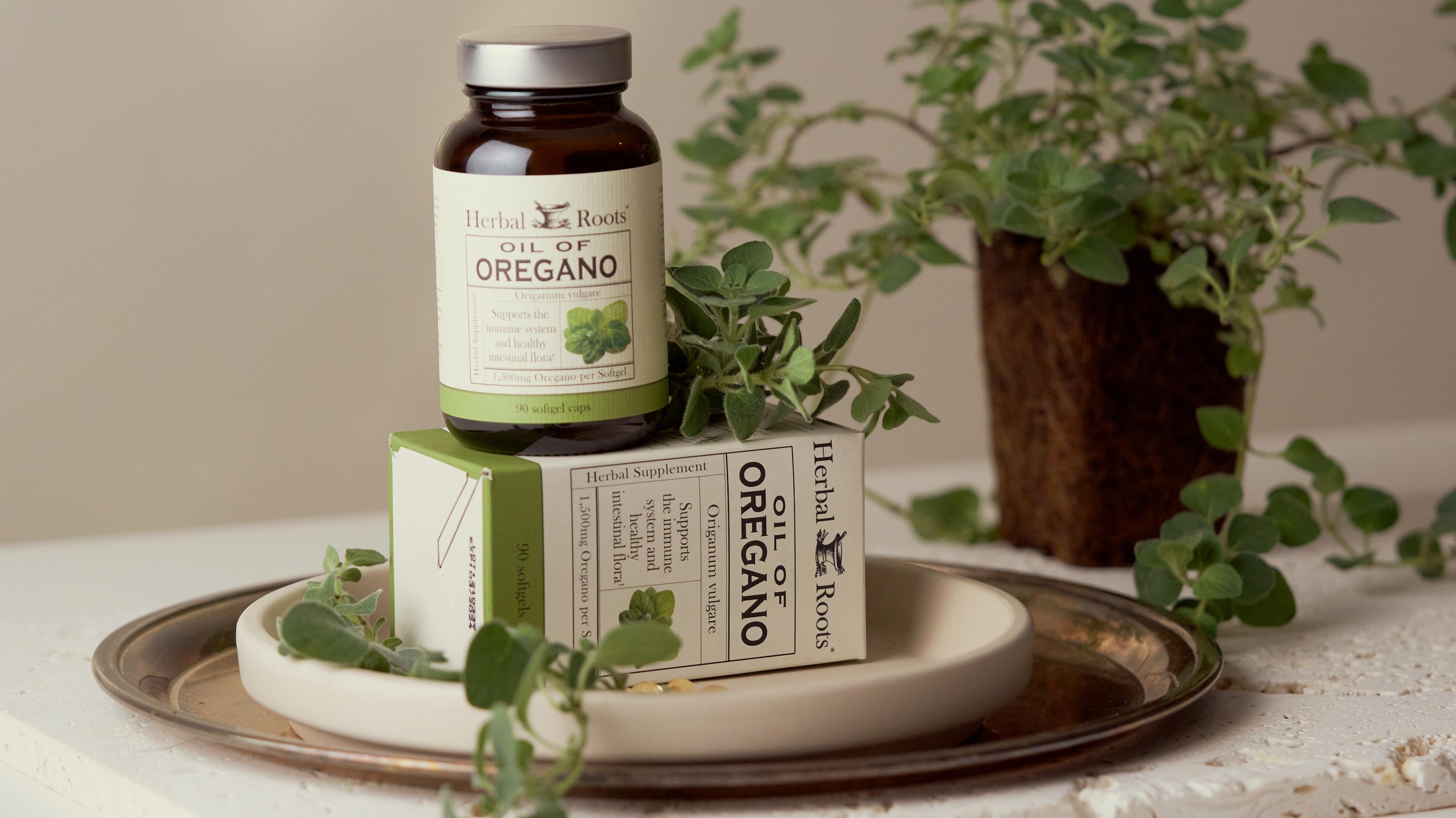 A slab of porous stone cutting board with a brass plate on it. On the brass plate is a ceramic plate with the box of Herbal Roots oil of oregano laying on its side. On top of the box is a bottle of Herbal Roots Oil of Oregano, matching with the box a green and white label. In the background in an oregano plant.