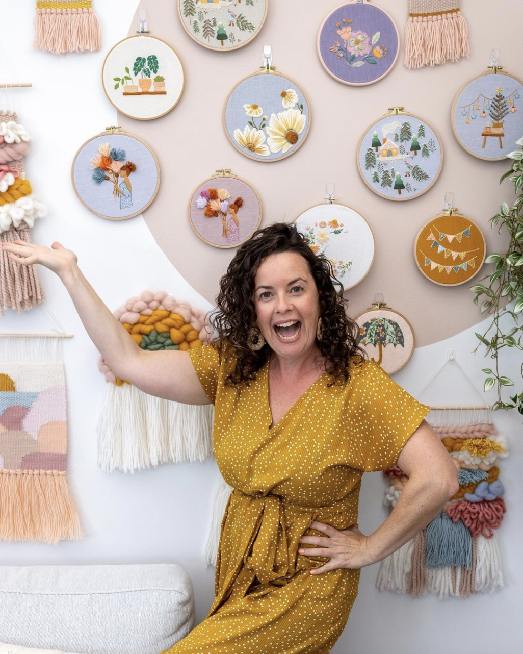 This is a picture of Clever Poppy creator Julie, gesturing to all her embroidery creations hanging on the wall.