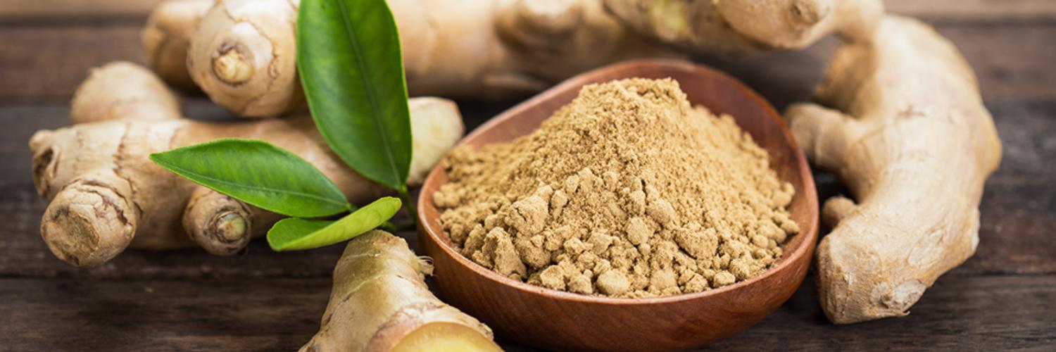 Raw ginger on table near a wooden bowl with powdered, brown ginger in it and leaves on a wooden table