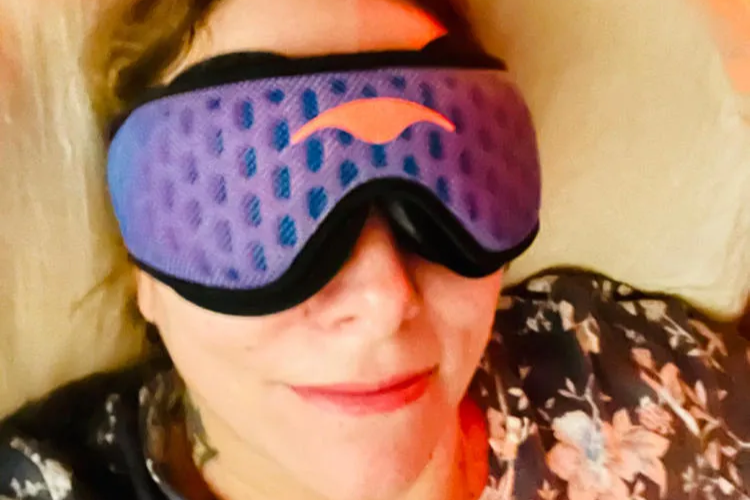 A smiling woman wearing a blue mesh eye mask that benefits side sleepers.