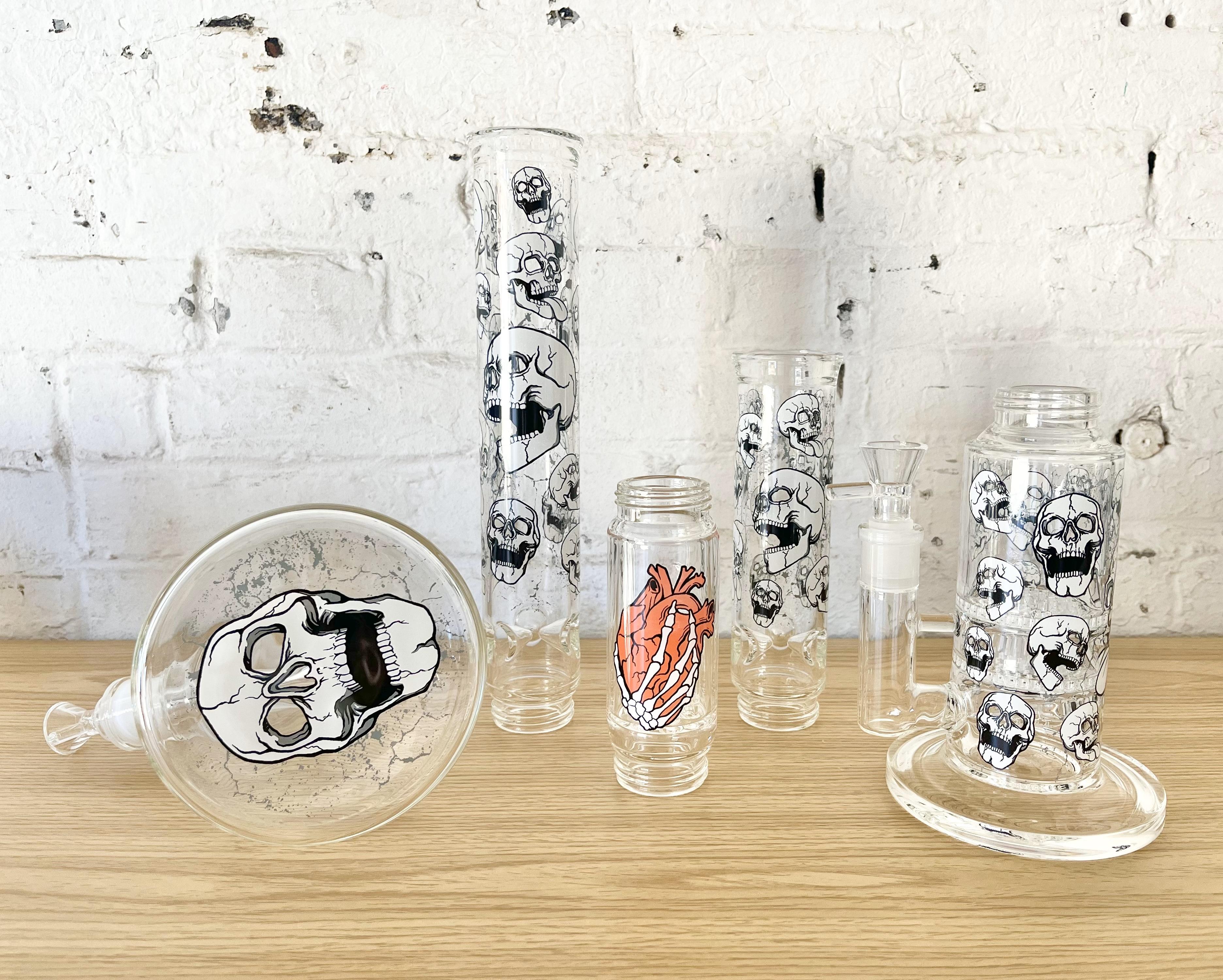 Disassembled clear Prism modular components, including a beaker base, tall mouthpiece, and percolators, all decorated with the Tommy Devoid Skelton Quotes decal. The designs feature distressed skulls and a skeleton hand gripping a heart, showcasing the morbid and ghastly collection theme on individual glass parts.