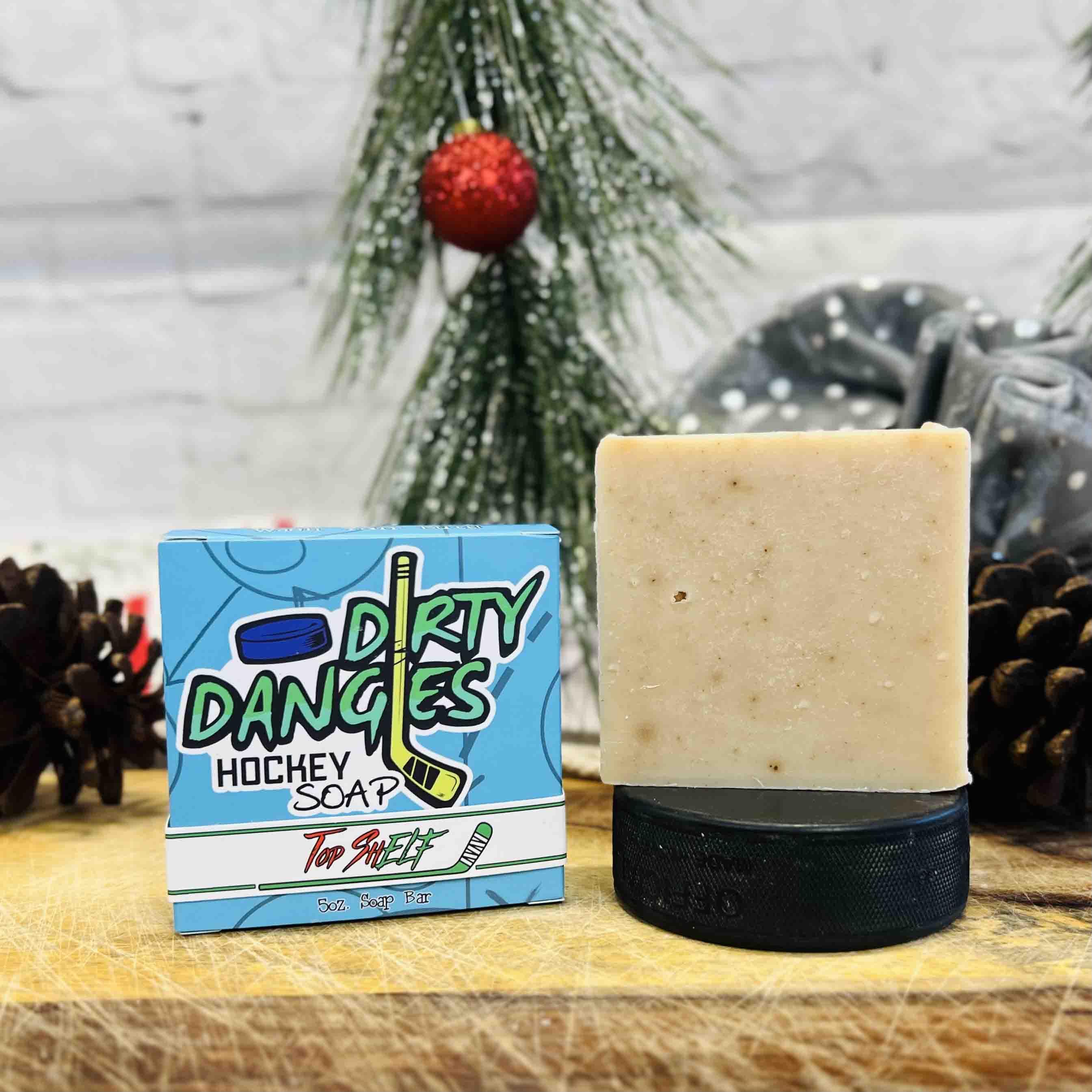 A blue box of Top ShELF hockey soap and a tan soap bar on top of a hockey puck on a christmas background. Dirty Dangles Holiday hockey soap.