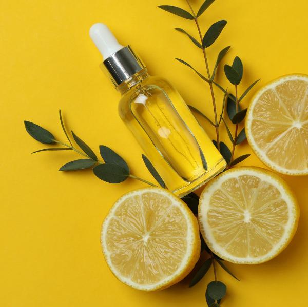 yellow background. clear essential oils dropper surrounded by green plants and three lemon slices.