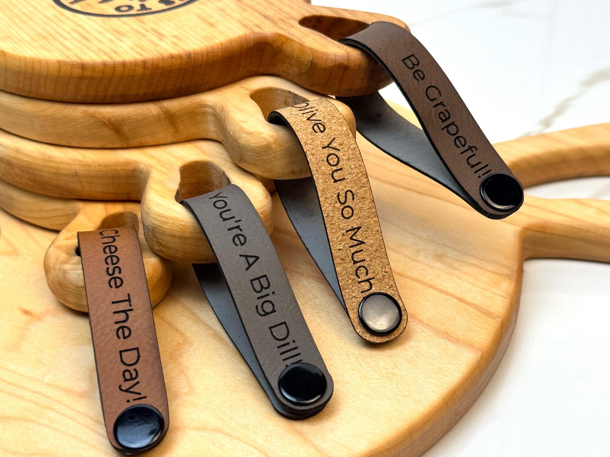 Maple wooden coaster single serve mini charcuterie board set with vegan leather straps engraved with puns: Be Grapeful!!, Olive You So Much, You're A Big Dill!, and Cheese The Day! Coasters have a natural finish.