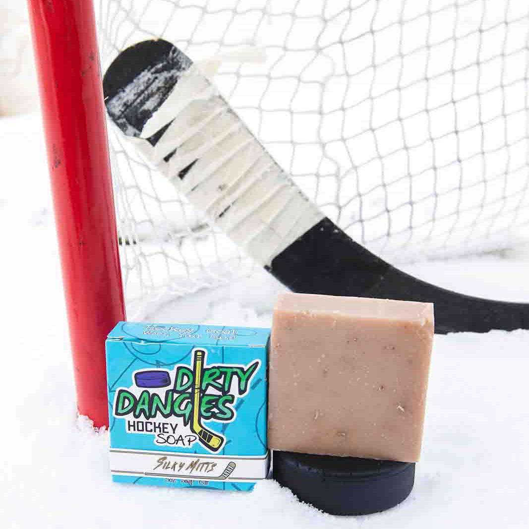 A black soap bar sits in the snow with a hockey goal, hockey puck and a hockey stick. Puck Drop