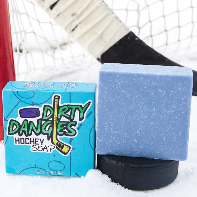 A blue bar of dirty dangles hockey soap sits on a hockey puck in the snow with a hockey stick and a hockey goal. The Michigan Scent
