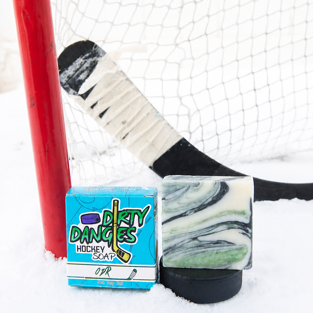 A handmade soap bar with a green and white marble pattern sits on a black hockey puck next to its blue and white 'Dirty Dangles Hockey Soap' box, set in the snow in front of a hockey goal and stick.