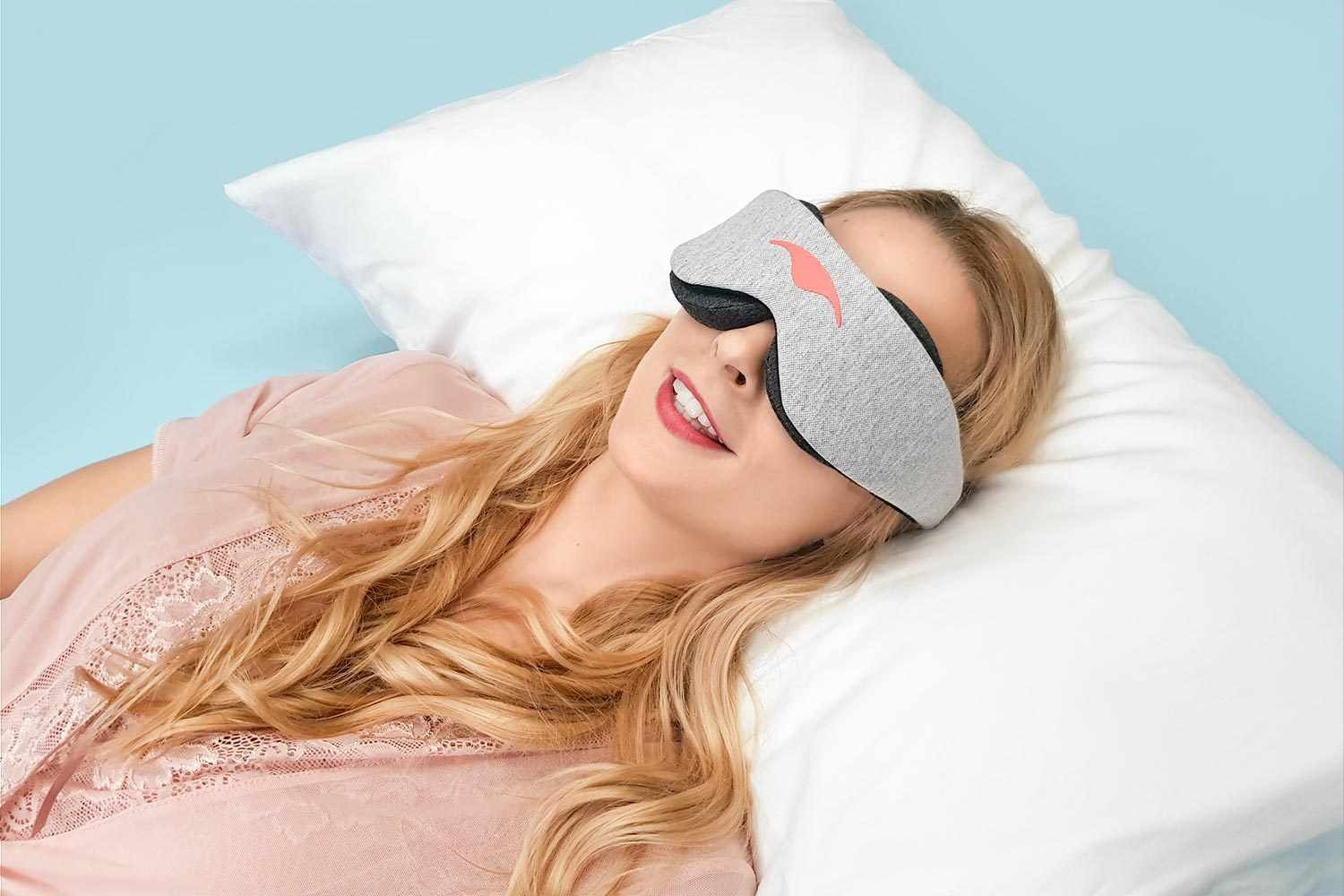 A smiling blonde girl lying down on a pillow and wearing one of the best blackout sleep masks from Manta Sleep.