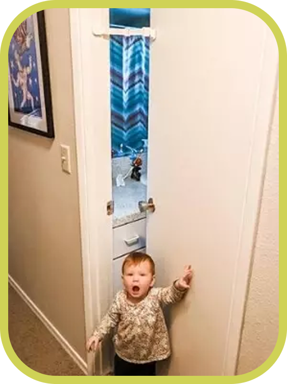 A toddler stands in a hallway near a door secured with The Door Buddy door strap. The strap baby proofs the doorway by keeping the opening small enough to help prevent toddlers from entering.