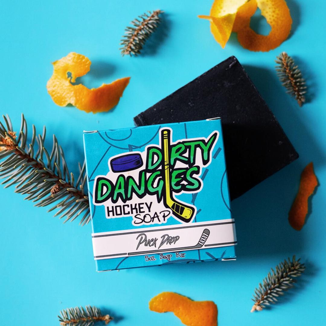 A bar of dirty dangles hockey soap puck drop scent on a blue background with pine and orange peel.