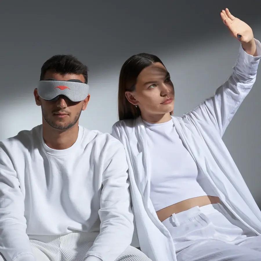 A man wearing a sleep mask for shift work sleep disorder. A girl is seated beside him blocking light with her hand.